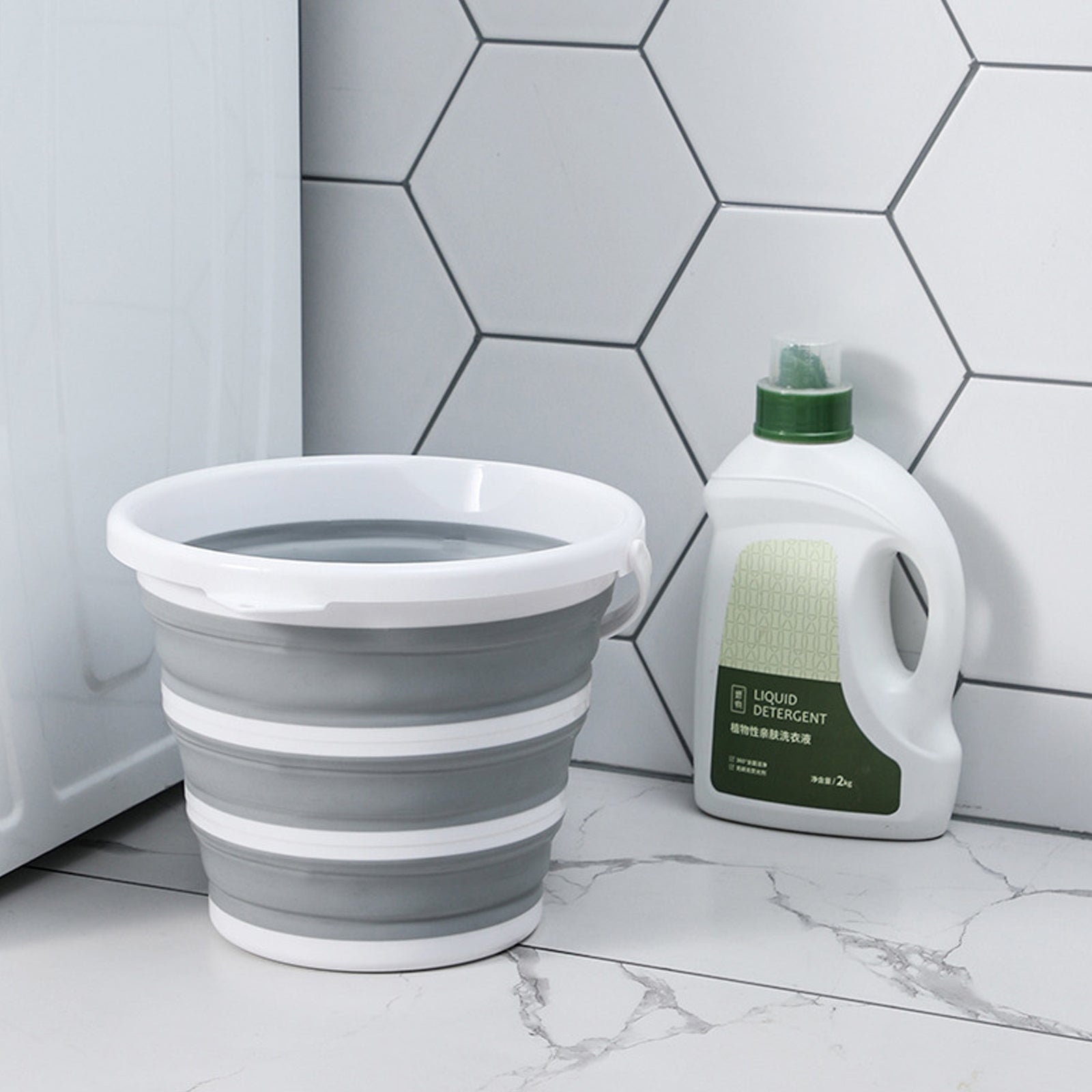 Collapsible gray bucket and liquid detergent bottle on a bathroom floor with tiled walls.