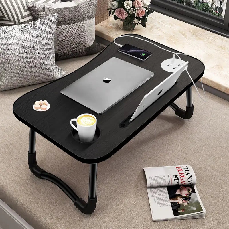 Black adjustable laptop desk on a carpeted floor with a laptop, cup, and magazine.